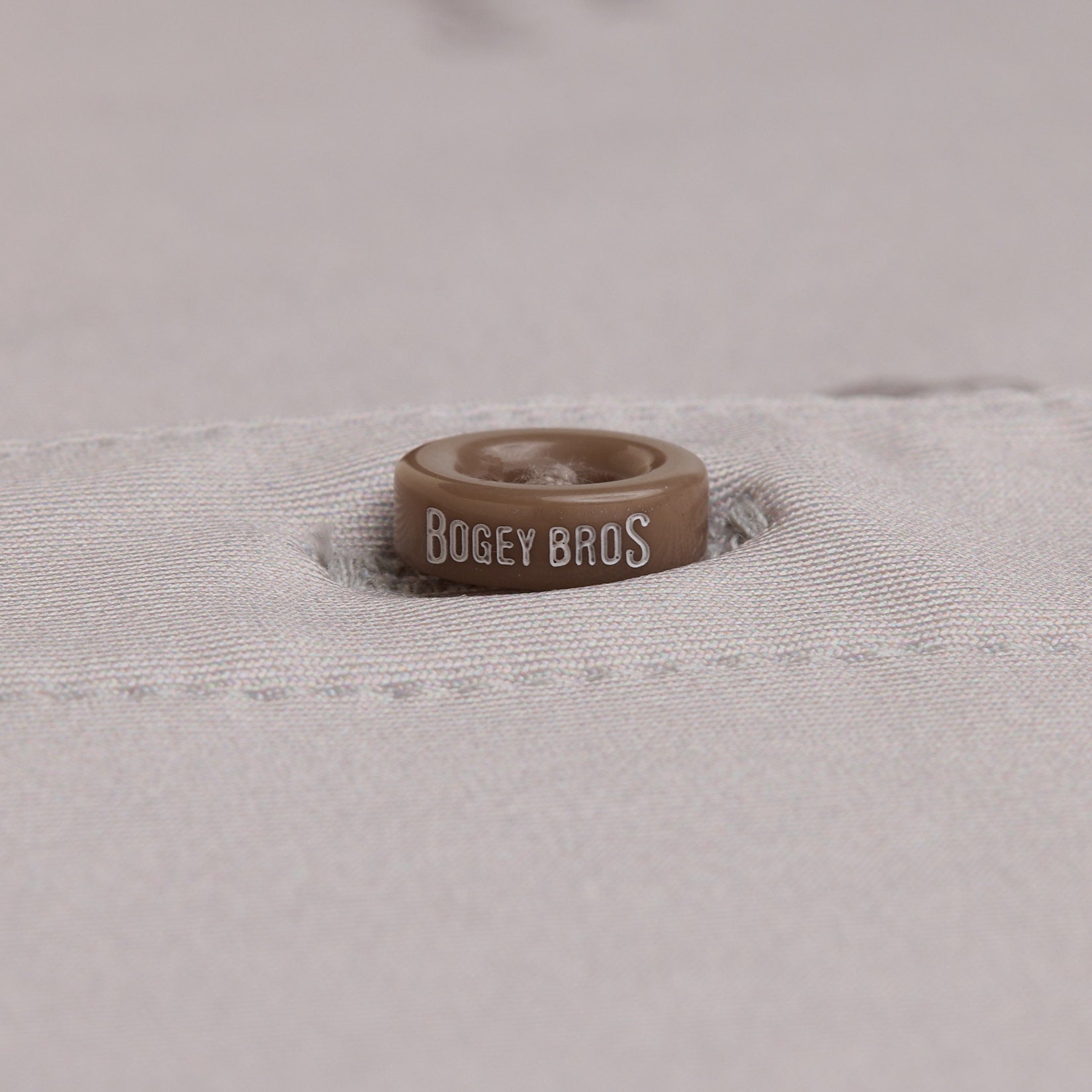 A close-up of a brown button with BOGEY BROS in white letters, sewn onto a light gray fabric. The button is partially embedded in a stitched buttonhole, and the fabric texture is clearly visible.