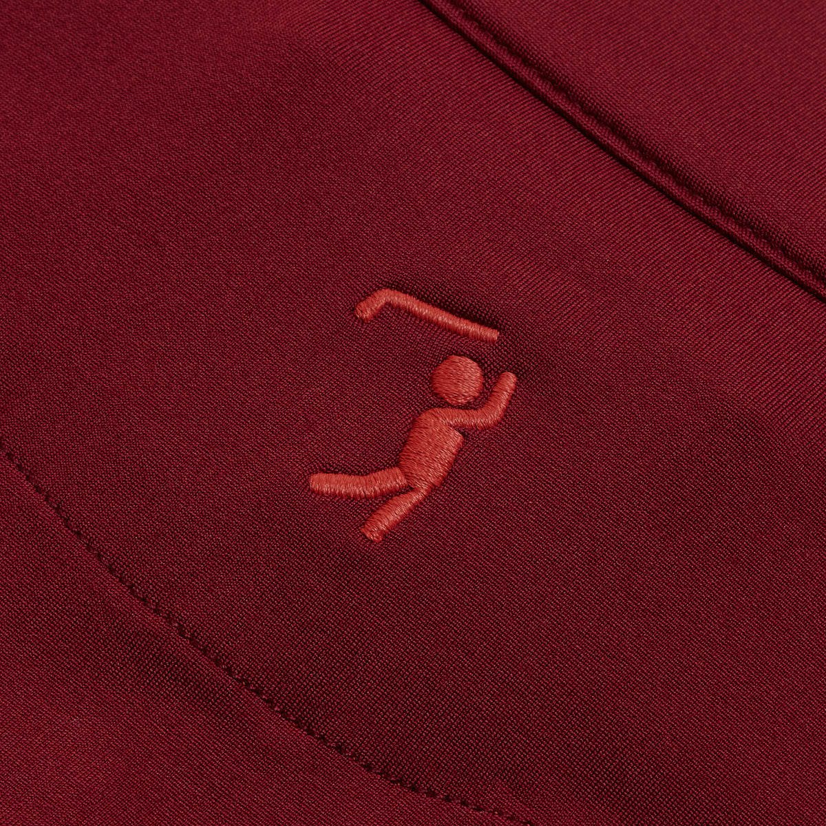 Close-up of the Burgundy - Q-Zip’s maroon fabric, featuring a small, embroidered figure swinging a golf club overhead in stitching that is a slightly lighter red than the base. The smooth background displays a diagonal seam behind the embroidery. Engineered with moisture-wicking properties for all-day comfort, this piece seamlessly blends performance and style.