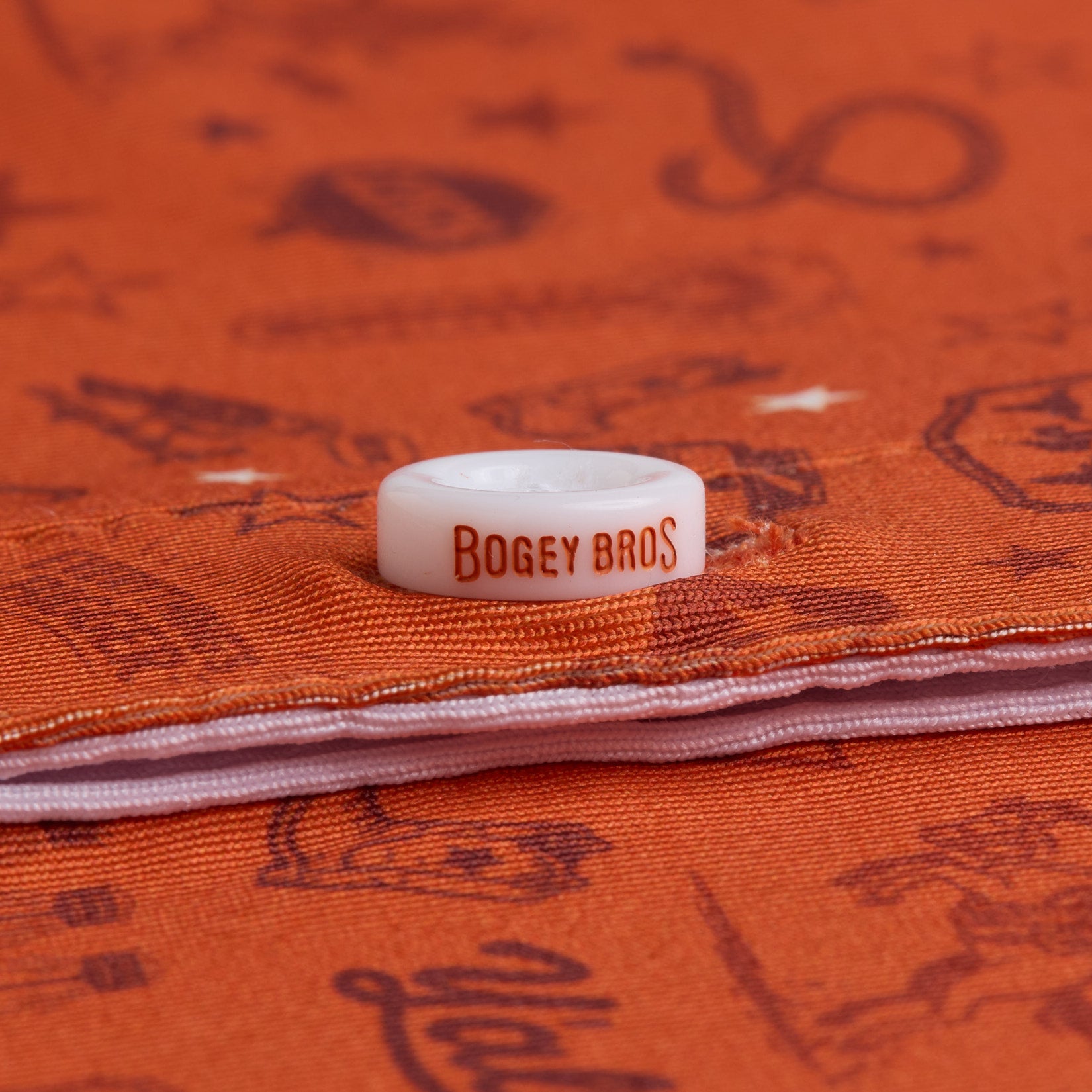 A white golf ball marker with BOGEY BROS in orange text rests on orange fabric with dark brown illustrations, including animals, objects, and shapes. A white zipper runs horizontally beneath the marker.