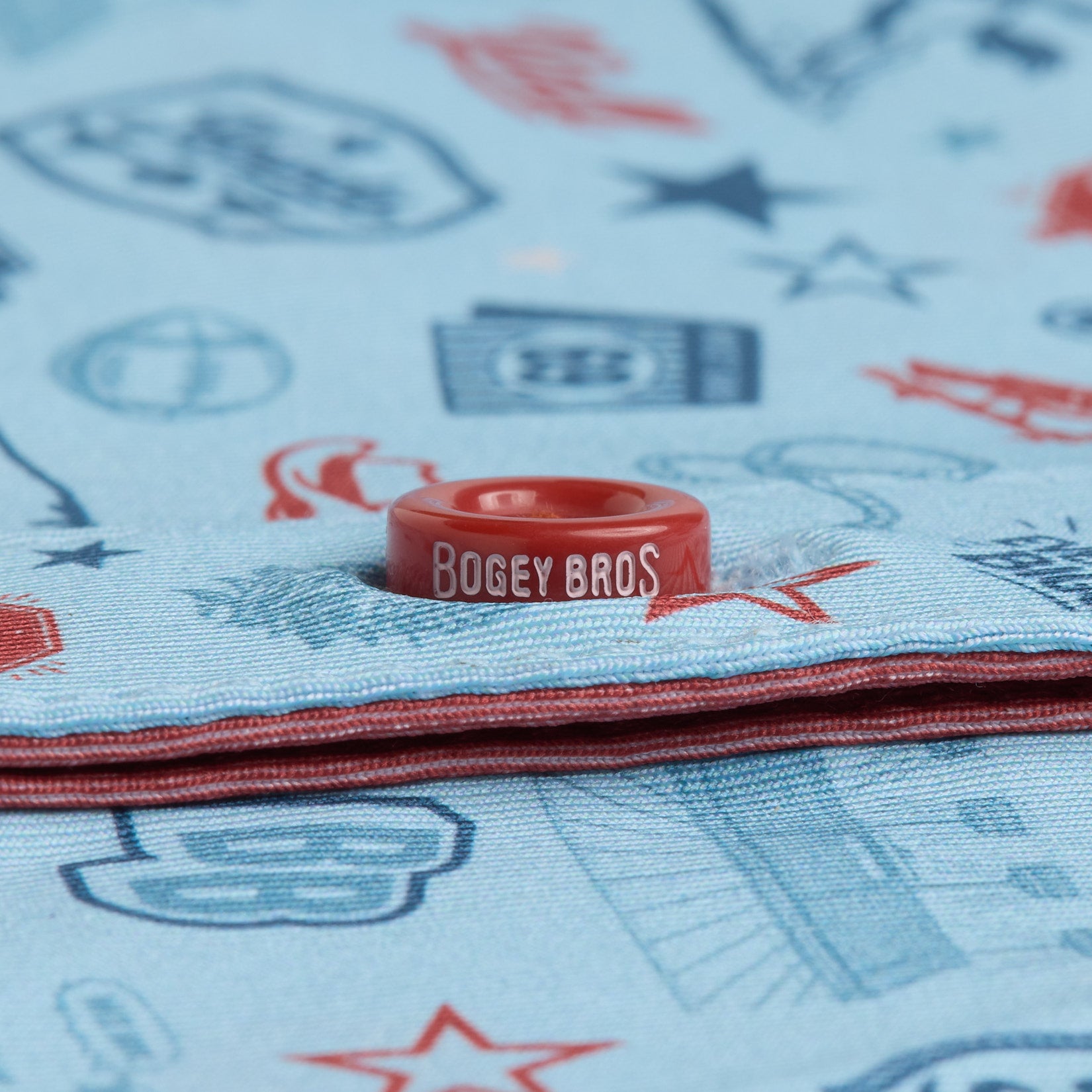 A red plastic marker labeled BOGEY BROS sits atop light blue fabric with red and blue golf-themed prints, including stars and golf balls. The fabric is folded, showing layered edges.