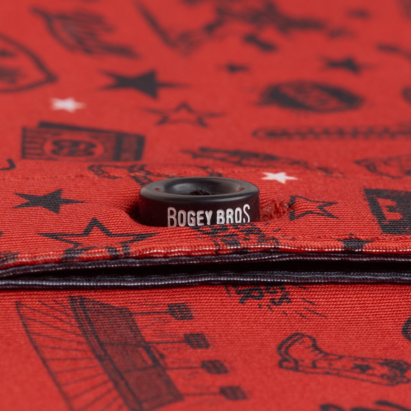 Close-up of a red fabric featuring black, sports-themed illustrations and stars. A black snap button in the center has “BOGEY BROS” written in white, standing out against the patterned background. The fabric’s edge and stitching are visible at the bottom.