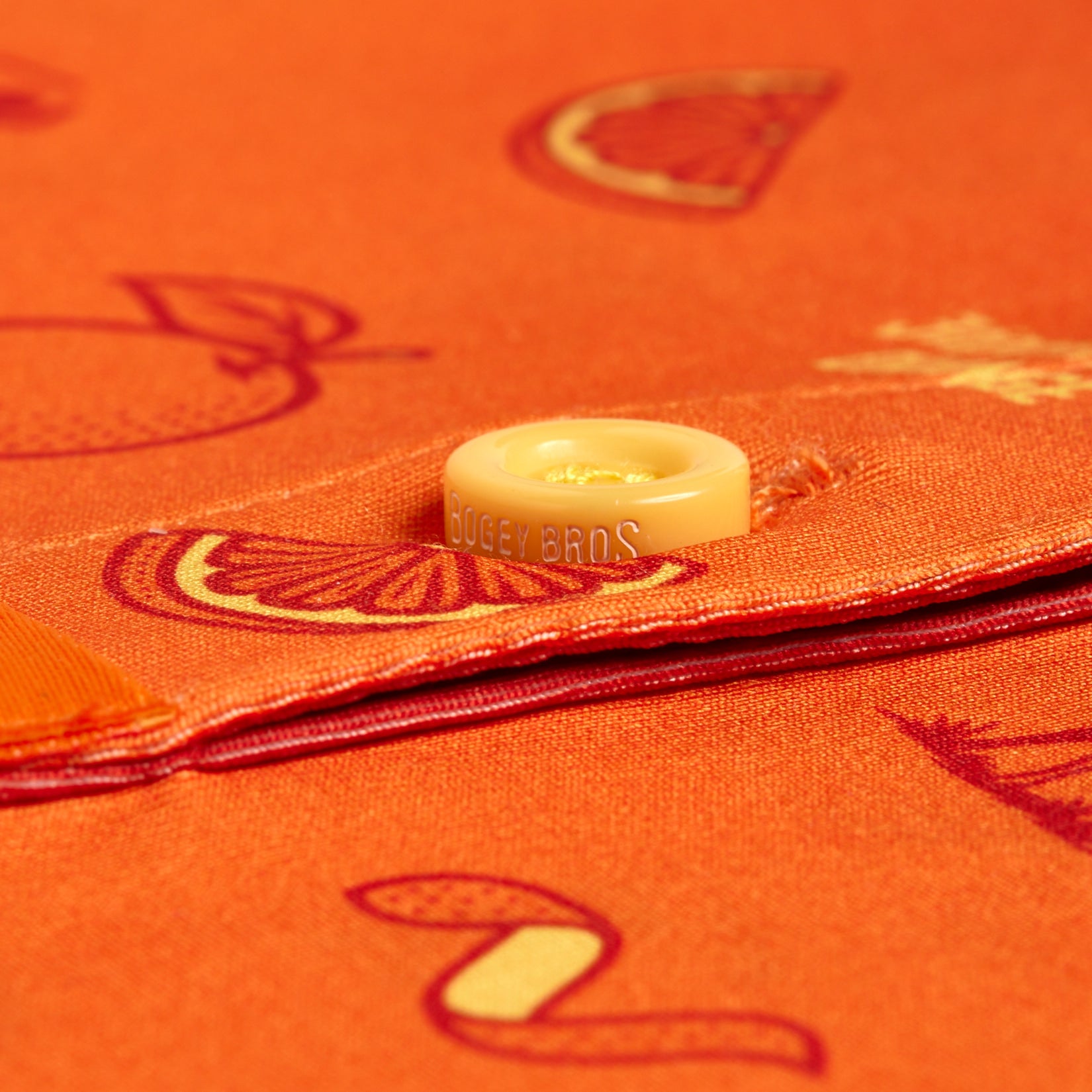 Close-up of Main Squeeze - Women's Polo in orange, featuring red fruit and worm illustrations. A yellow “BOGEY BROS” button accents the stitched edge, showcasing its playful pattern and moisture-wicking fabric.