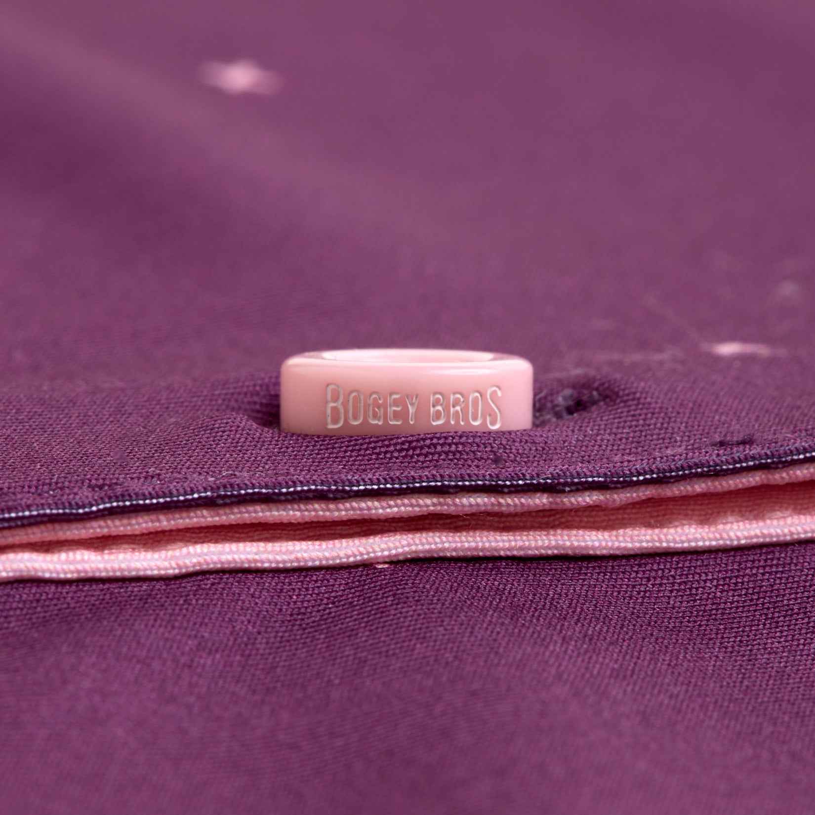 A close-up of the Exes - Plum - Women's Polo shows a pink button engraved with BOGEY BROS, sewn onto purple fabric with light pink stitching and visible layers, designed for an athletic fit.