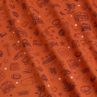 A burnt orange fabric features Texas-themed illustrations: cowboy hats, boots, cacti, stars, armadillos, stadiums, footballs, the word “Howdy,” lassos, the state outline, and the phrase Everythings Bigger, all in a dark shade, scattered across the material.