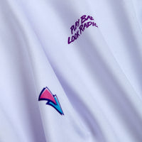 A close-up of the Chillax - Polo in white athletic fabric shows purple “PLAY BAD LOOK RADICAL” text and a colorful geometric arrow in blue, pink, and purple. The smooth, moisture-wicking material displays gentle folds.