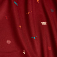 A close-up of the Desert - Polo shows maroon fabric with turquoise and yellow cacti, longhorn skulls, circles, and white HOOEY BROS text. The slightly textured fabric offers moisture wicking for comfort.