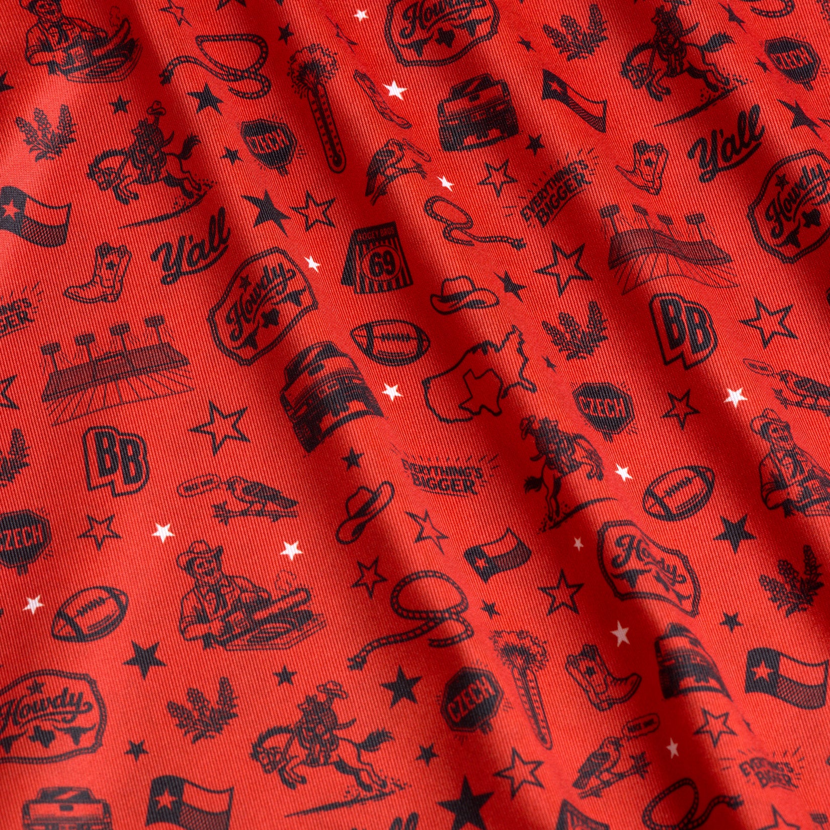 Red fabric with black illustrations: cowboy boots, hats, guitars, Texas state outline, cacti, sheriff badges, stars, speech bubbles with “Y’all,” Texas flags, boots, and Howdy phrases, all scattered and slightly wrinkled.
