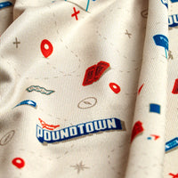 A close-up of the Poundtown - Polo's beige fabric, showcasing playful graphics—lips, speech bubbles, geometric shapes, and bold blue “POWDTOWN” text—in red, blue, and gray accents on a moisture-wicking textured surface.