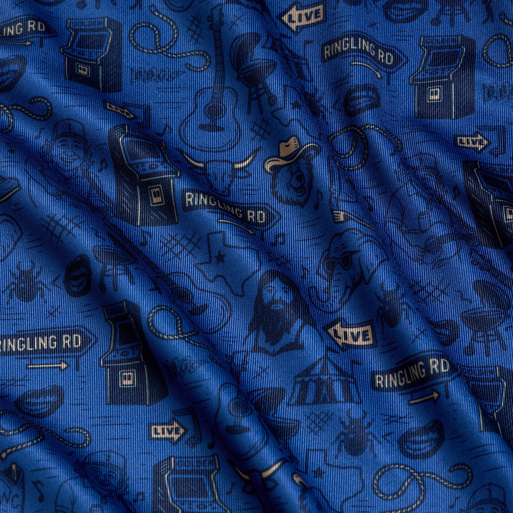 Ringling Road - Polo features gently rippled blue fabric adorned with playful black outlined illustrations, including guitars, cowboy hats, microphones, arcade machines, stars, a bearded man, and “Ringling Rd” street signs. The textured surface adds dimension and subtle shadows to the lively pattern.