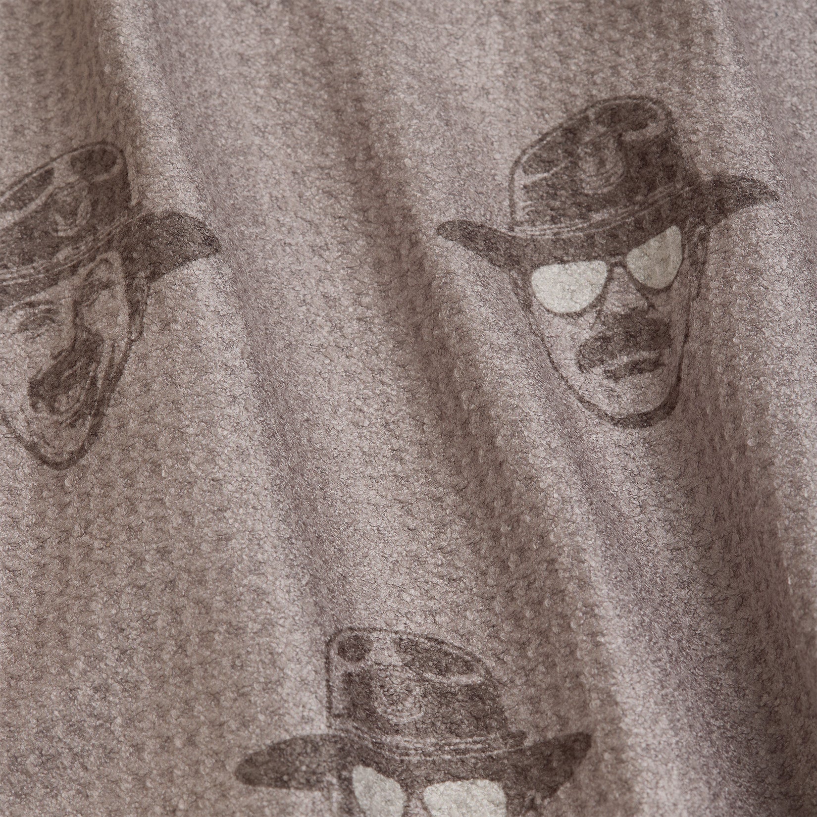 Wrinkled taupe fabric with a waffle texture features a repeating print of a mans face wearing a cowboy hat and sunglasses. The illustrated faces are dark brown, with some showing mustaches, and are spaced evenly across the material.