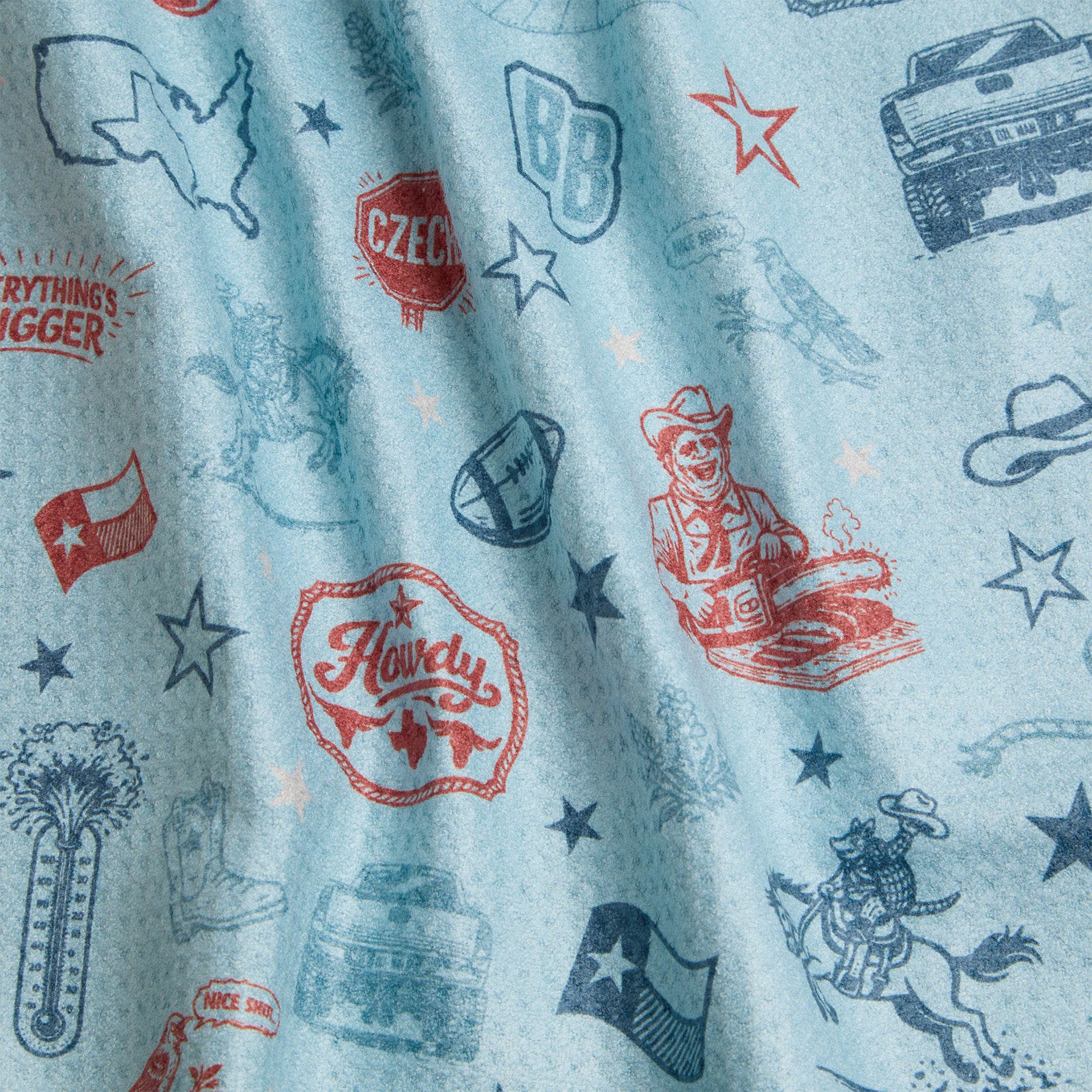 Light blue fabric with various red and blue Texas-themed illustrations, including a cowboy, a truck, cowboy hats, the word “Howdy,” the Texas state outline, a football, stars, and other Western motifs scattered throughout. Some areas show shadowed folds.