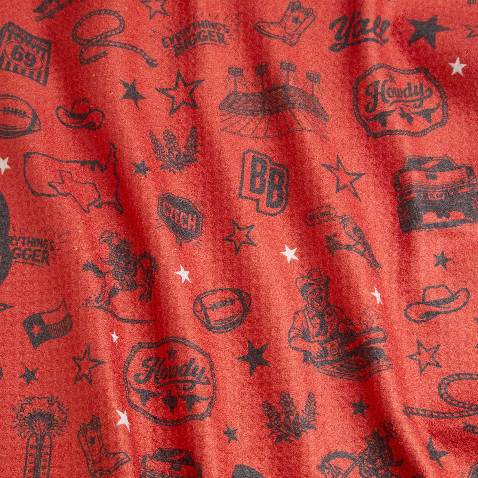 A red fabric printed with various black Western-themed icons, including cowboy boots, hats, stars, stadium, bandana, bolo tie, a football, state of Texas, the word Howdy, a truck, Yall, and Everythings Bigger. The fabric is slightly wrinkled.