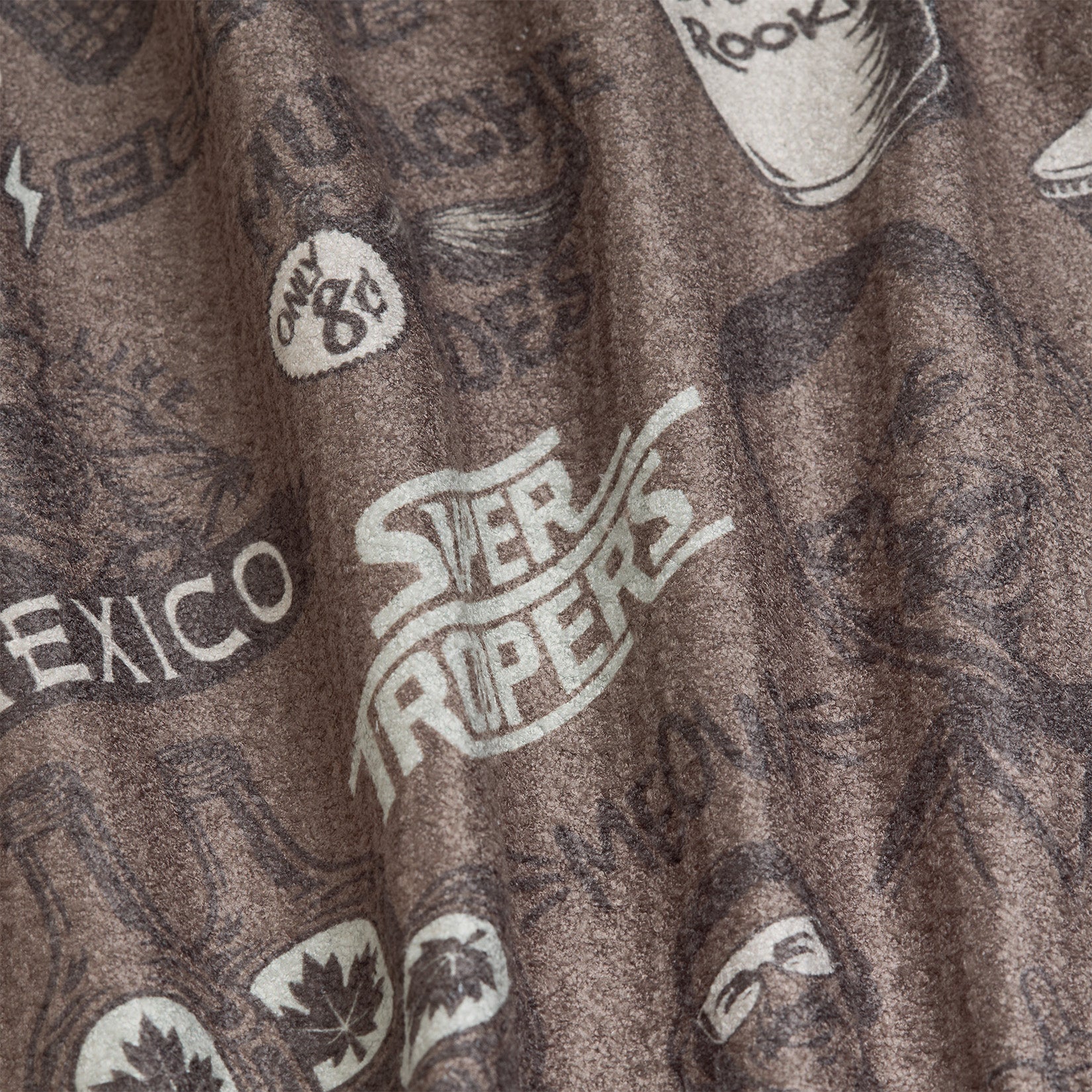 A brown fabric with white printed graphics, including the words SUPER TROOPERS, a Route 66 sign, bottles, a map of Mexico, a cup with ROOKS, and maple leaves; material appears wrinkled.