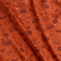 Orange fabric with repeating maroon Texas-themed illustrations, including cowboy hats, boots, armadillos, stars, footballs, BBQ grills, cacti, and words like “Howdy,” “Y’all,” and “Everything’s Bigger.” The fabric is lightly wrinkled.