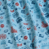 Light blue fabric with dark blue and red Texas-themed illustrations, including cowboy hats, boots, cacti, stars, maps, “Howdy,” “Y’all,” and “Everything’s Bigger” text. The fabric appears slightly wrinkled, adding texture to the pattern.