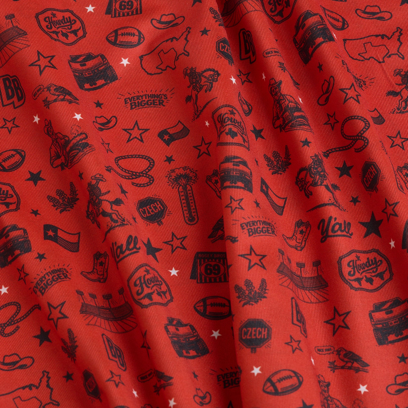 Red fabric printed with small black illustrations and text, including cowboy hats, Texas shapes, boots, footballs, stars, “Howdy,” “Y’all,” “Everything’s Bigger,” and other Western-themed motifs. The fabric has visible folds and shadows.