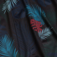 A close-up of the Haolie Nights - Women's Polo shows teal palm fronds and a red monstera leaf on dark fabric with subtle shadowy leaves. Slight gathering adds texture, and the polo features UPF 50 protection.