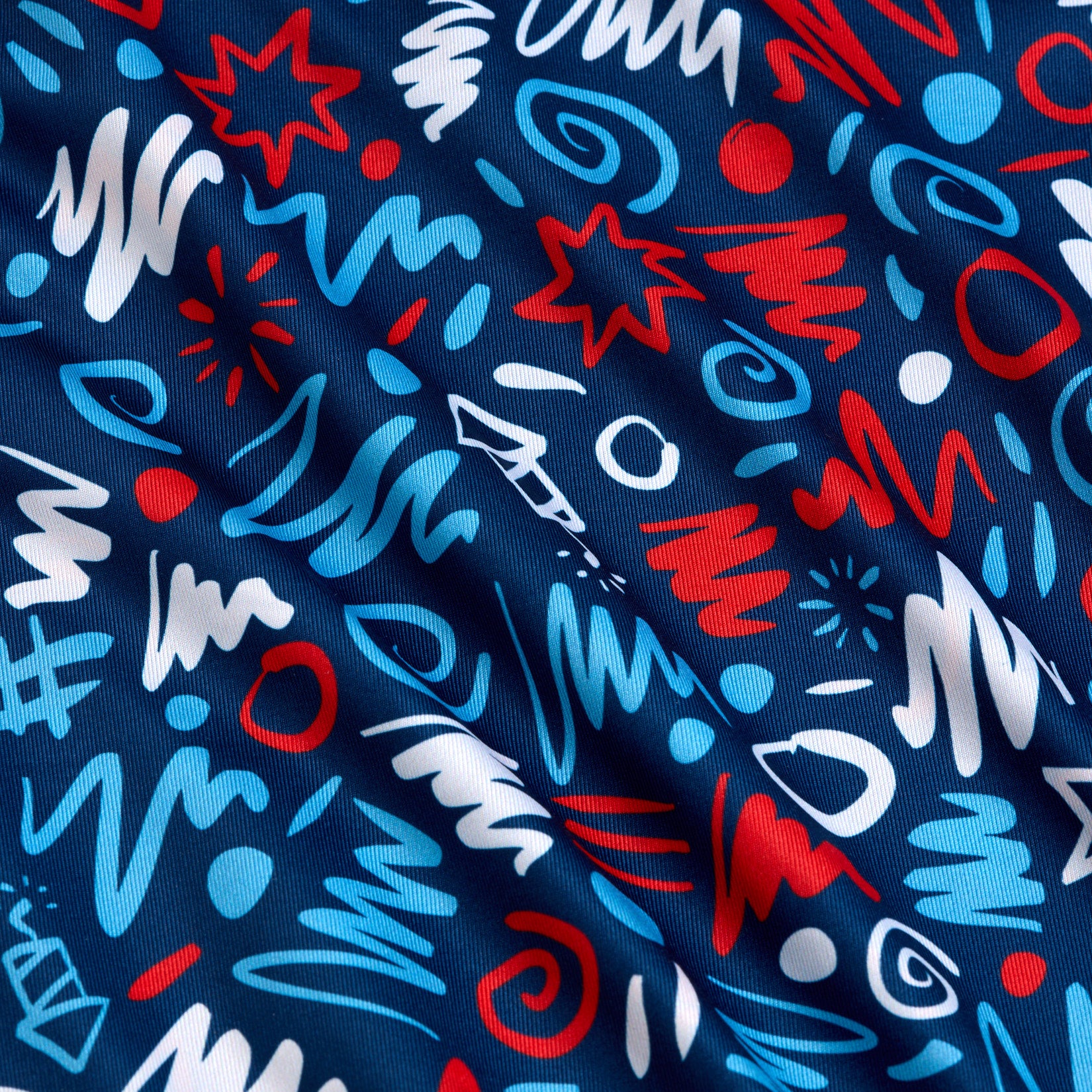 Blue fabric with a playful, abstract pattern featuring squiggles, zigzags, swirls, stars, and dots in red, white, and light blue. The fabric has subtle folds and a slightly shiny texture, creating a sense of movement across the surface.