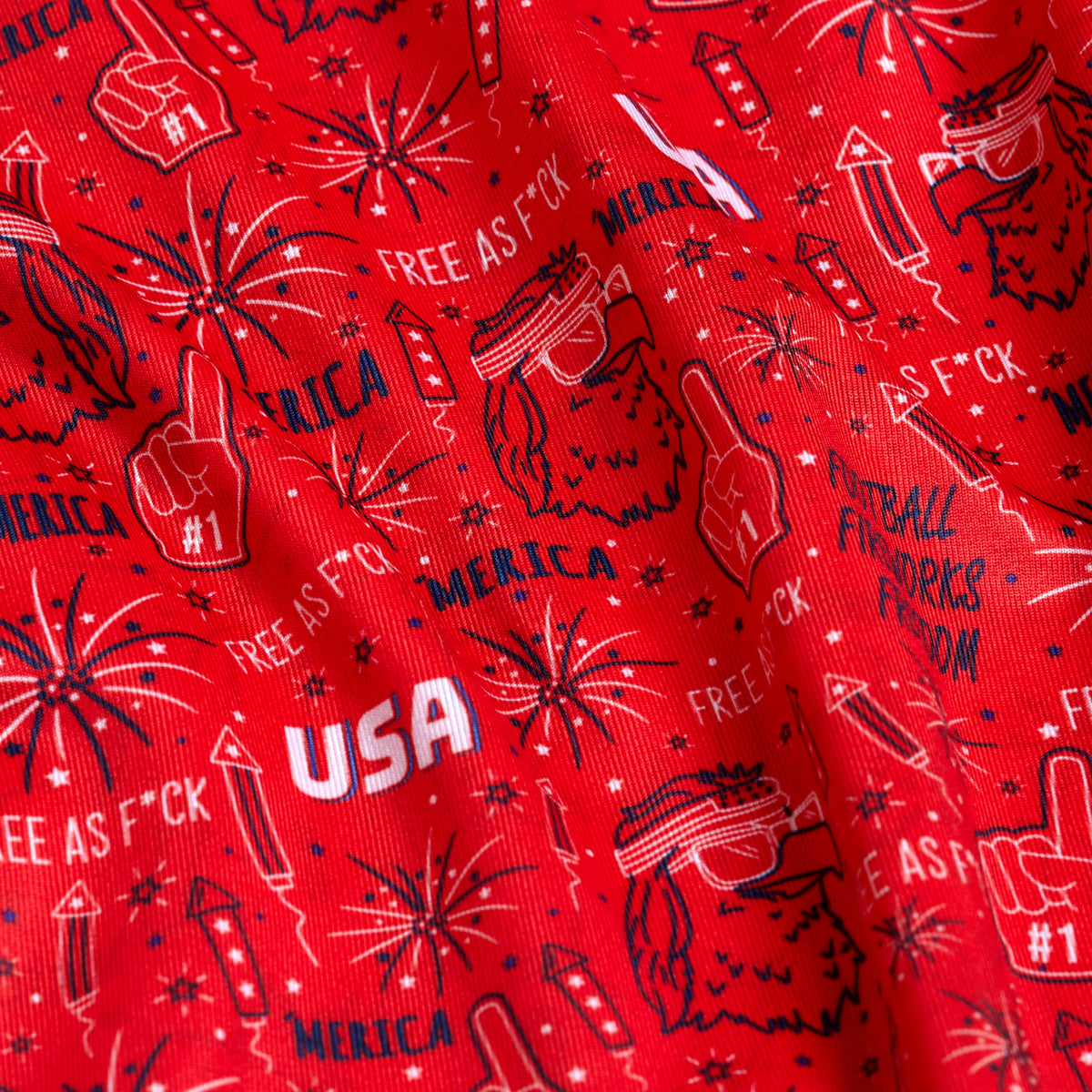 A close-up of 'Merica - Red - Polo: red fabric with white and blue patriotic designs—fireworks, rockets, #1 foam fingers, sunglasses, bold phrases—and UPF 50 protection for sun safety.