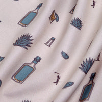 Close-up of the Reposado - Women's Polo fabric with playful blue and brown bottles, agave plants, lime wedges, salt shakers, and cocktail glasses on a light beige background. Soft, textured fabric offers UPF 50 sun protection.