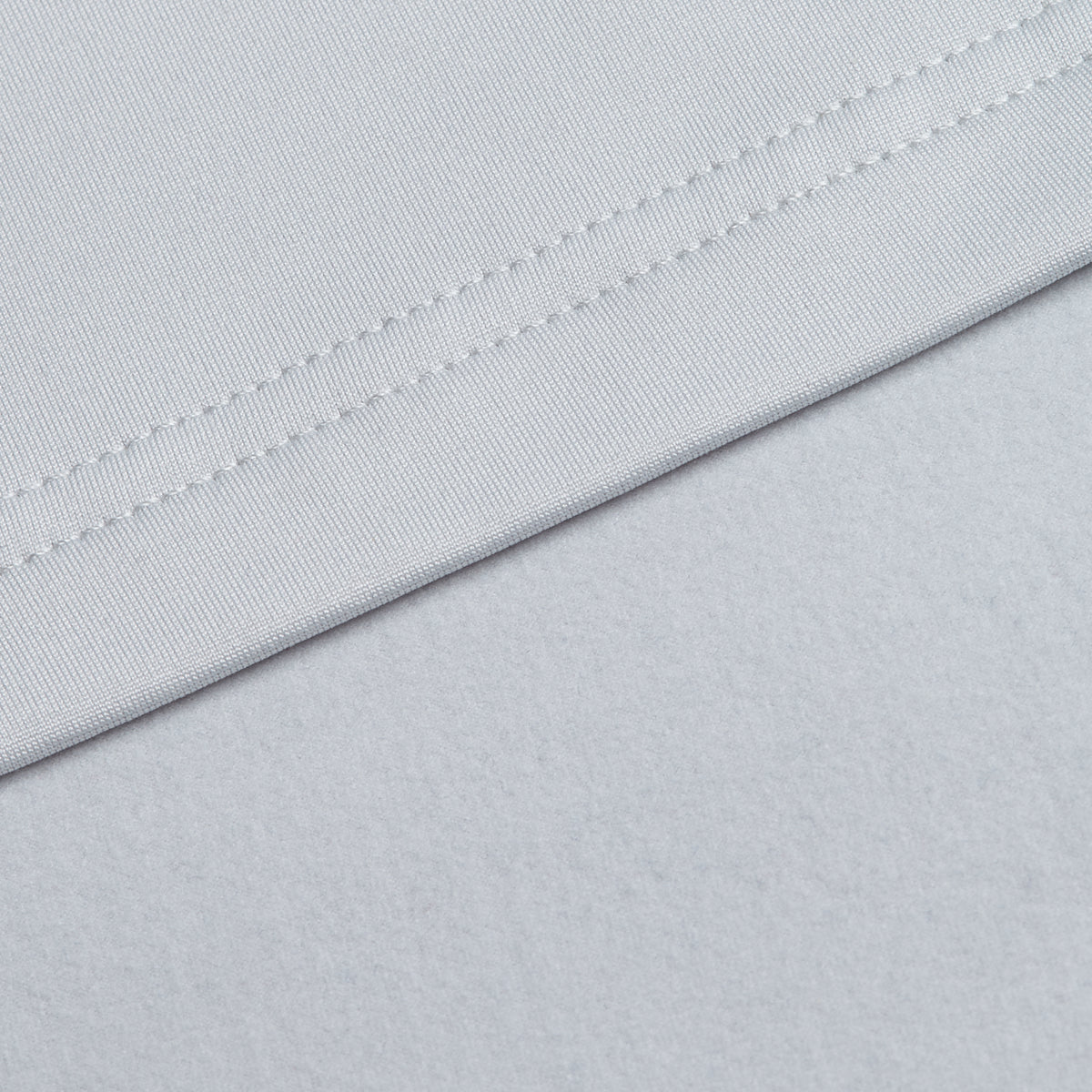 Close-up of two light gray fabric surfaces from the Bone - Hoodie. The upper fabric, which features visible stitching along its edge and a smooth finish, is commonly chosen for athletic wear due to its moisture-wicking properties. It is laid diagonally across the softer, more matte lower fabric.