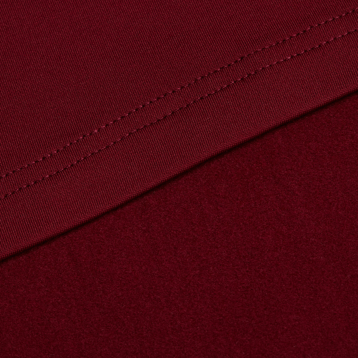 Close-up of a burgundy fabric from the Burgundy - Hoodie, featuring a visible seam running diagonally across the material. The upper section displays fine stitching, while the lower portion appears smooth, showcasing the texture and rich color characteristic of this premium hoodie. The fabric is free from patterns or additional elements, emphasizing its high quality and subtle moisture-wicking properties.