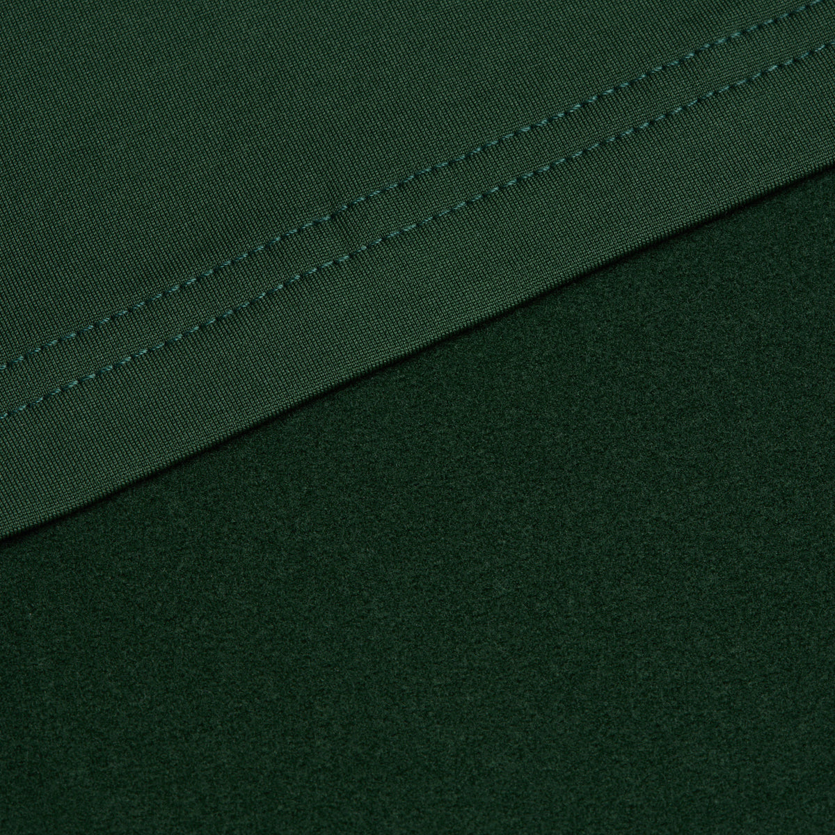 A close-up of the Forest Green Hoodie shows dark green fabric with visible stitching along the edge. One section overlaps another at a diagonal, highlighting the textural contrast between two surfaces of the material. The neat, parallel stitching suggests a well-made garment crafted from stretchy, moisture-wicking fabric.