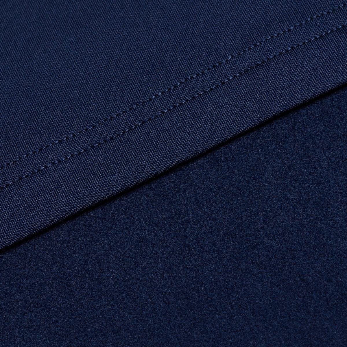 Close-up of two dark blue fabric surfaces from the Navy - Hoodie. The upper fabric is smooth with visible double-stitched seams, providing durability and support, while the lower fabric has a soft, slightly textured look typical of moisture-wicking materials. Arranged diagonally, the fabrics contrast in texture and stitching, emphasizing comfort features and UPF 50 protection designed into the hoodie.