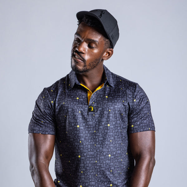 A muscular man in the Gusher - Polo, an athletic fit black patterned short-sleeve shirt with yellow accents, and a black cap tilts his head and smirks while posing against a plain light gray background.