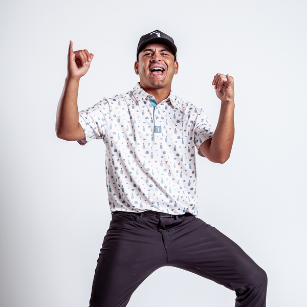 A young man in an athletic fit white Reposado - Polo with a pattern, dark pants, and a black cap poses energetically against a white background, smiling widely with both hands raised and fingers pointing up, full of excitement and confidence.