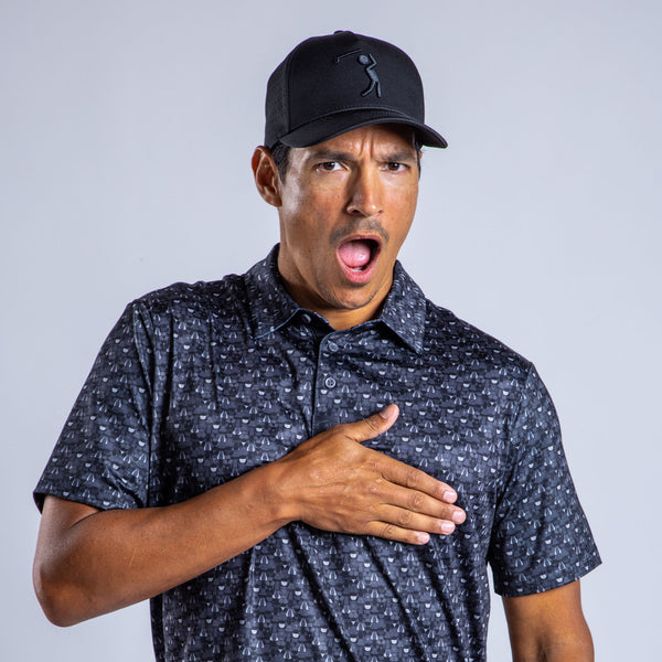 A man wearing the T&A - Fitty Shades - Polo with UPF 50 stands against a light gray background, surprised with mouth open and eyes wide, his right hand on his chest, and a black baseball cap on.