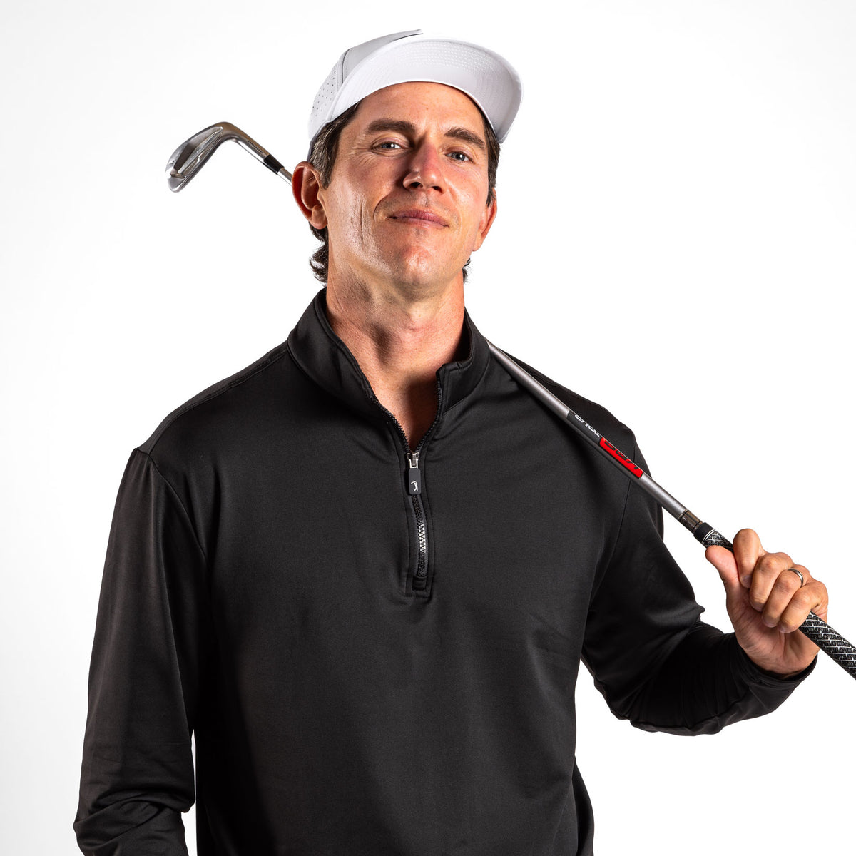 A man wearing the Black - Q-Zip with an athletic fit and a white cap stands confidently, holding a golf club across his shoulders with one hand. The background is plain white, and he has a slight smile on his face.