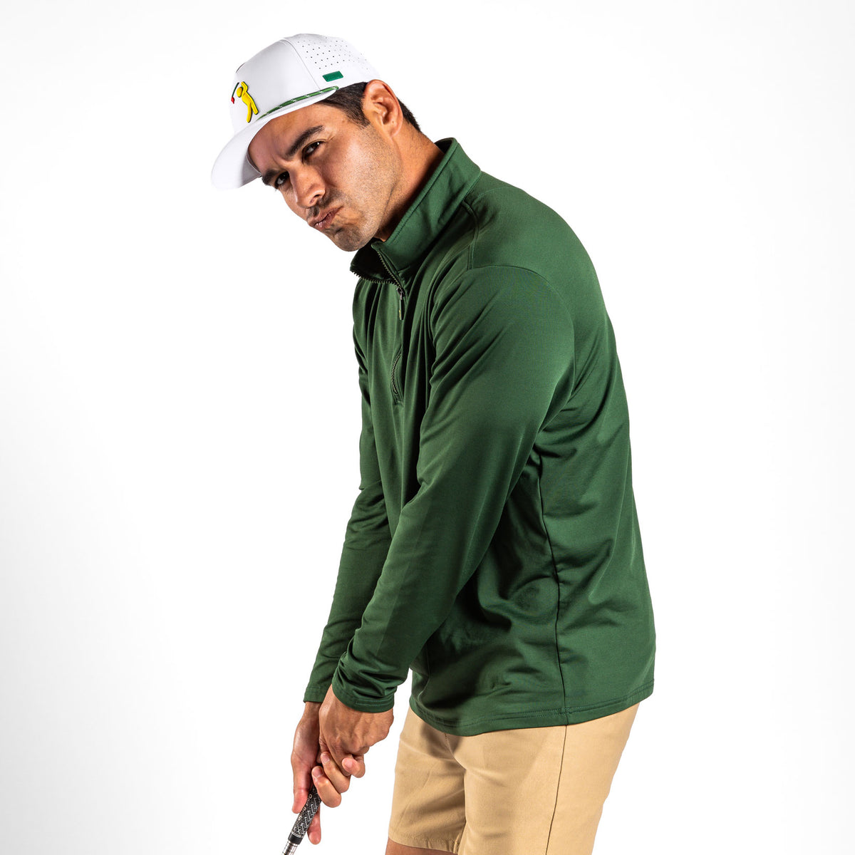 A man in a white golf cap, tan shorts, and the Forest Green - Q-Zip stands against a white background. Dressed in super stretchy, athletic fit apparel, he poses as if holding a golf club, looking directly at the camera with a playful yet intense expression and pursed lips.