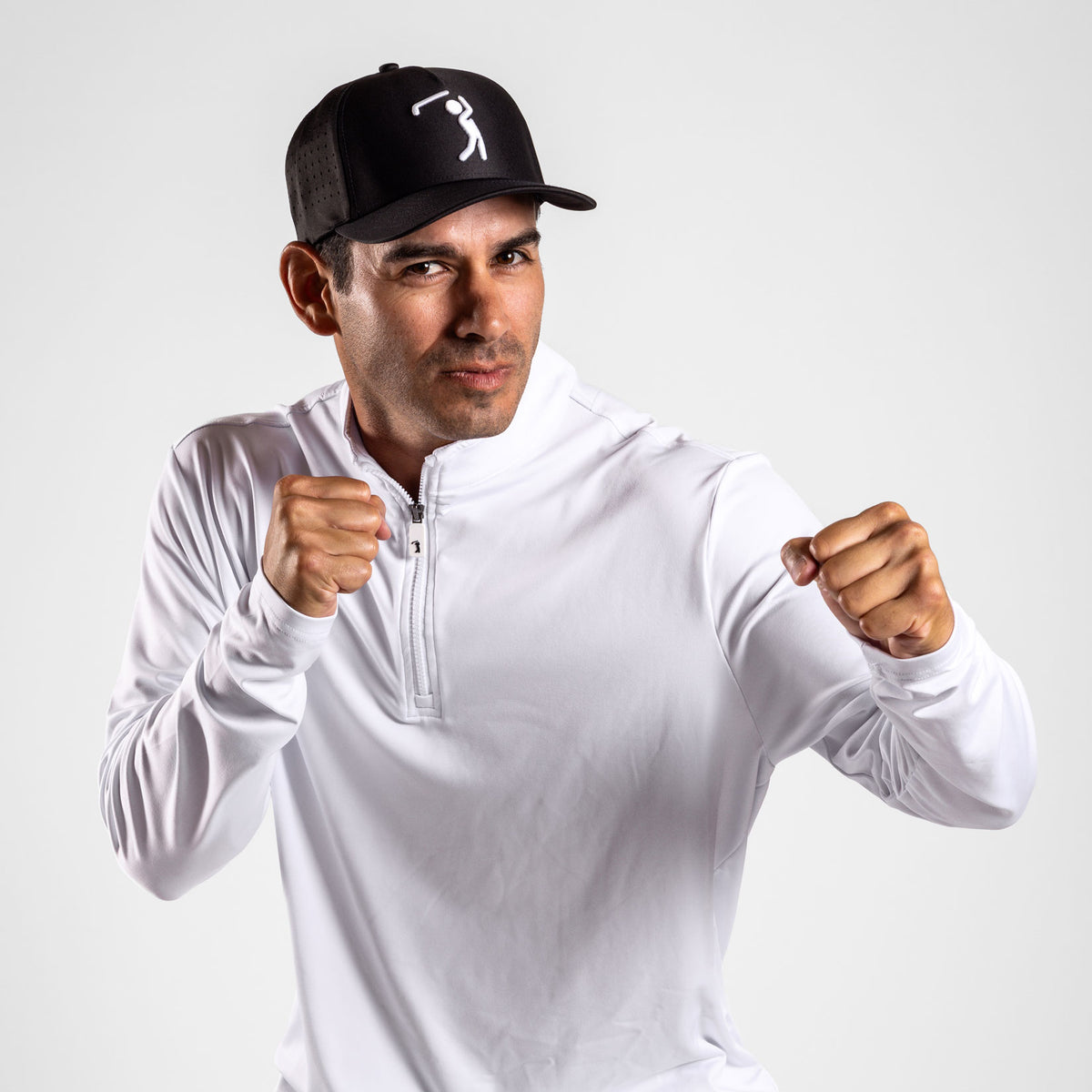 A man in a Black Cap featuring a simple white graphic of a swinging golfer and a White - Q-Zip with an athletic fit poses with fists raised as if boxing, exuding confidence and focus with a slight smirk. The plain light gray background emphasizes his stance. The White - Q-Zip provides UPF 50 protection, making it ideal for active days both indoors and outdoors.