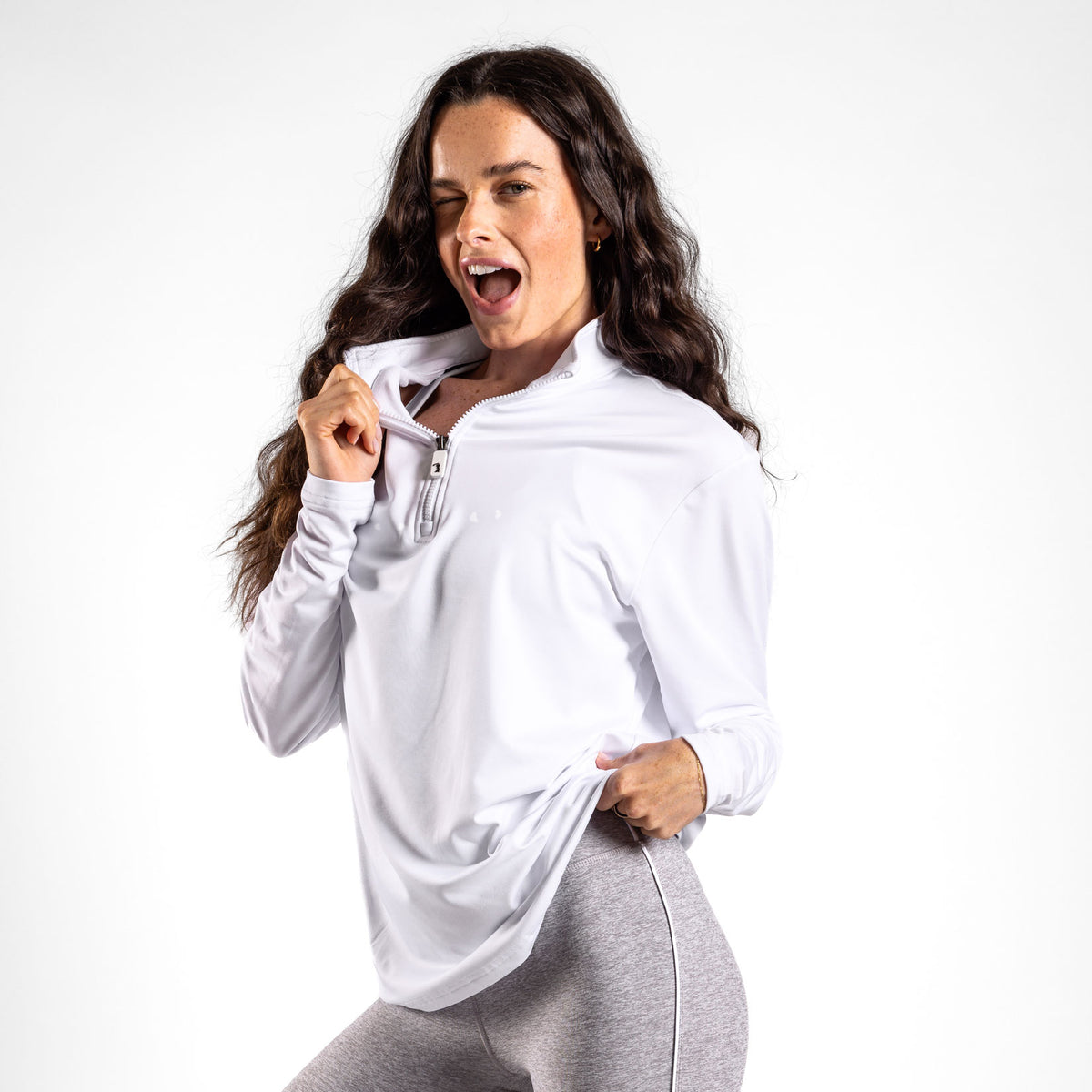 A woman with long, wavy brown hair stands against a white background, winking and smiling with her mouth open as she holds the collar and lifts the hem of her White - Q-Zip, which features an athletic fit, paired with gray leggings.