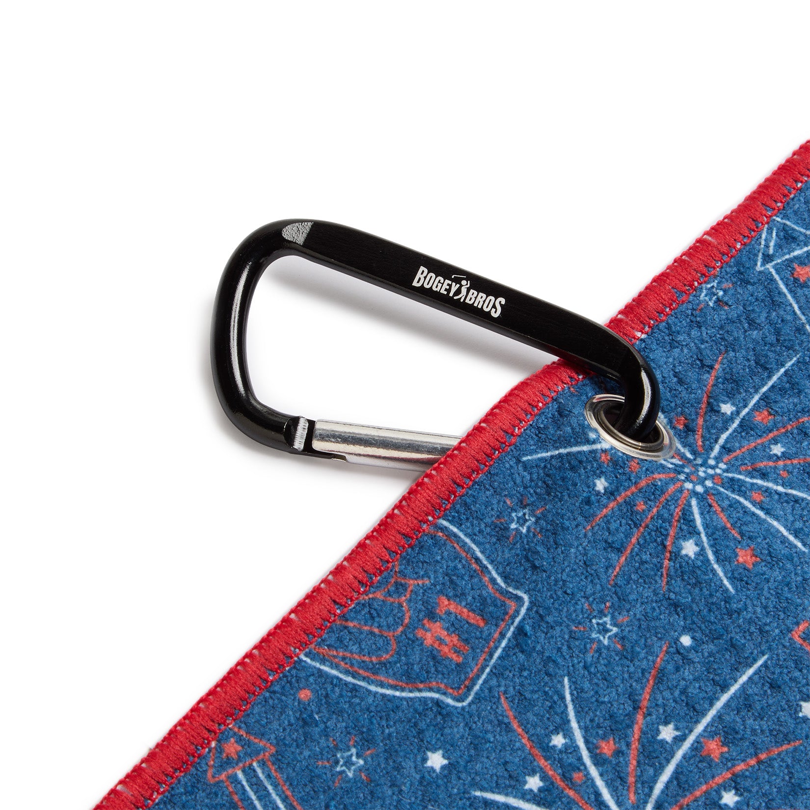 A black carabiner labeled BOGEY BROS is clipped to the metal grommet of a blue towel with red trim. The towel features white and red fireworks and festive patterns, including foam fingers and stars.