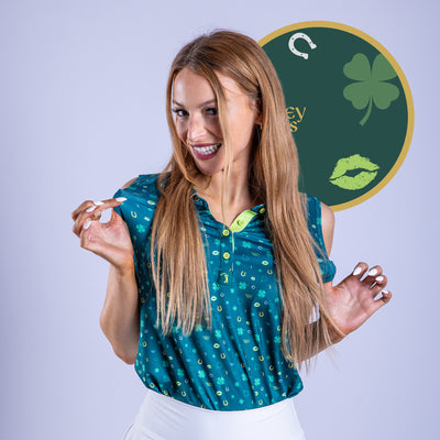 A smiling woman with long blonde hair wears the Gettin' Lucky - Women's Polo, a teal shamrock-patterned sleeveless top with UPF 50 and athletic fit, paired with a white skirt. She poses playfully in front of a green circle featuring lucky icons.