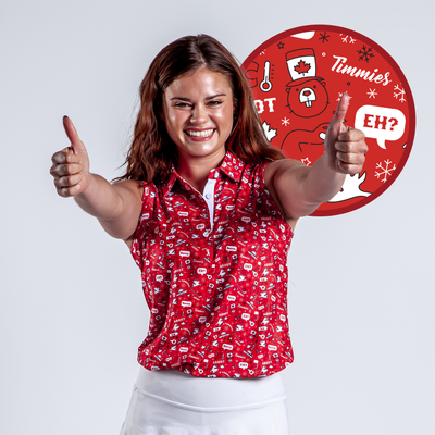 A young woman with long brown hair smiles and gives two thumbs up. She wears the O Canada - Women's Polo, a red patterned sleeveless shirt offering UPF 50 protection, along with a white skirt. A round graphic with Canadian symbols is behind her.