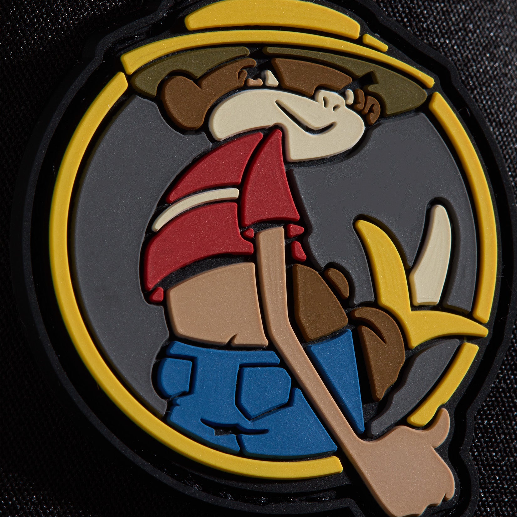 Cartoon monkey wearing a yellow hat, red crop top, and blue pants, holding a peeled banana and giving a thumbs-up. The design is outlined in yellow and black, set against a dark textured background.