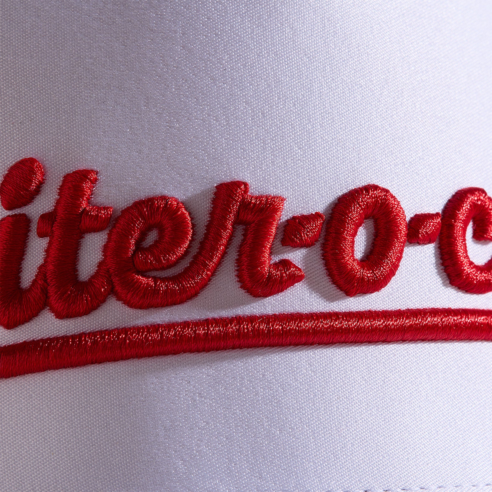 Close-up of red script embroidery on white fabric, showing the text iter-o- with bold, raised stitching. A red curved underline runs beneath the letters. The texture of the thread and fabric weave is clearly visible.