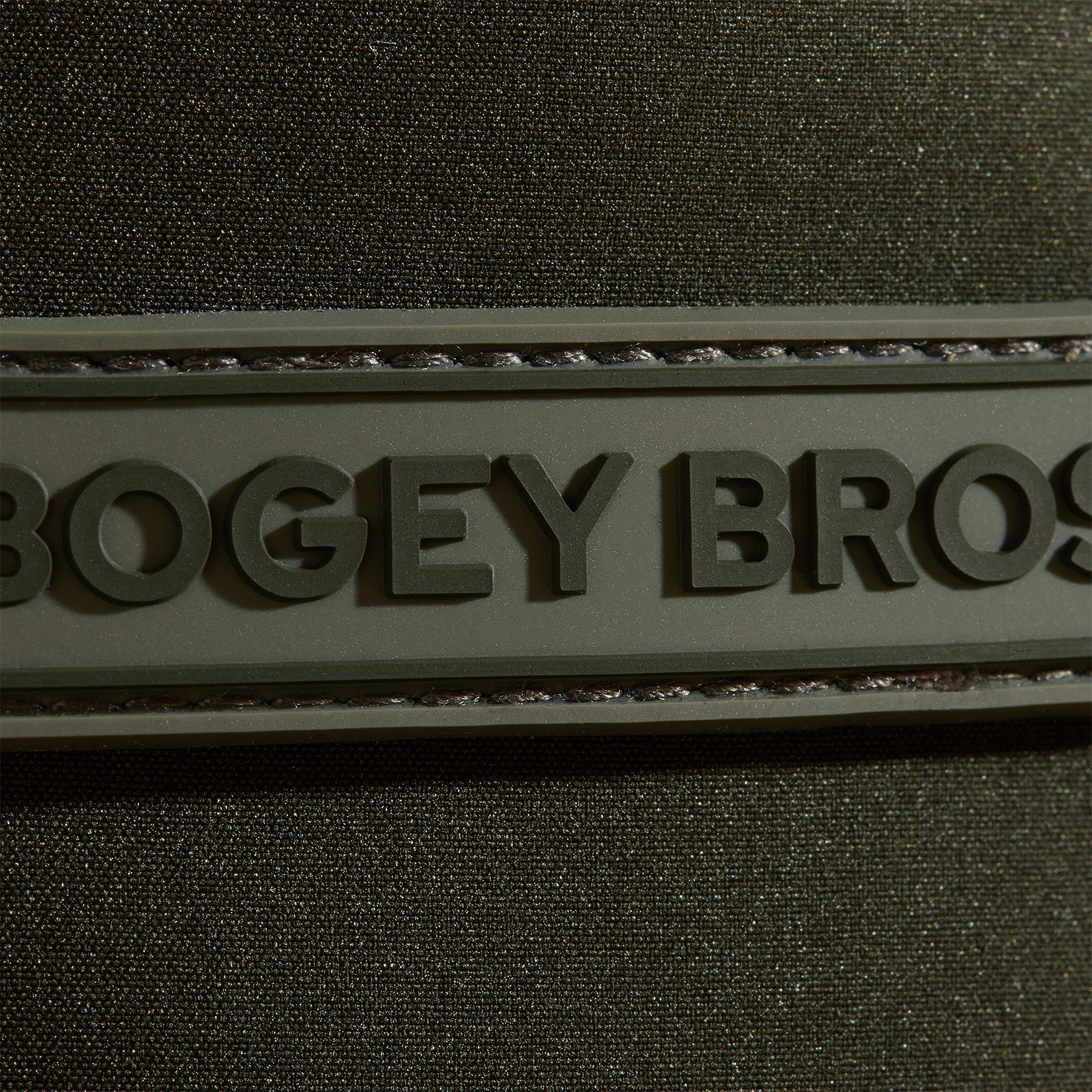 A close-up of a textured dark green fabric with a raised rubber patch that has the embossed, partially visible text BOGEY BROS. The patch is stitched onto the fabric, and the lighting highlights the materials texture and details.
