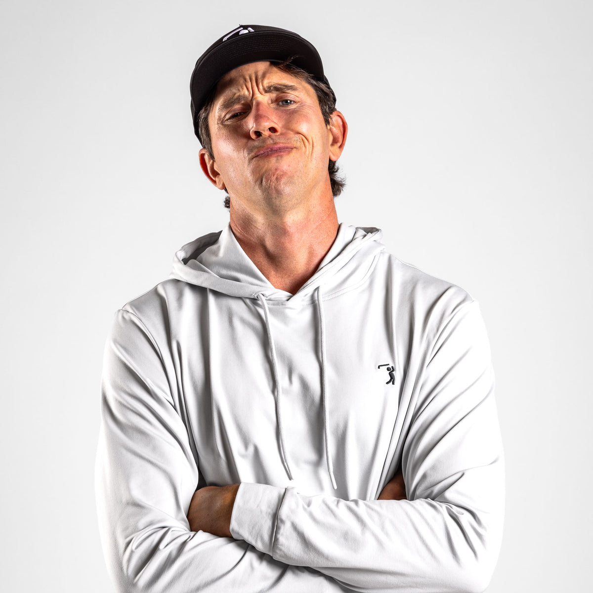 A man with a smirk and raised eyebrows stands against a light grey backdrop. He wears a black baseball cap and the Bone - Hoodie, a long-sleeved, light grey hooded shirt featuring a small golfer logo on the chest. Designed with UPF 50 protection for optimal sun safety without sacrificing style, he folds his arms across his chest and gazes directly into the camera.