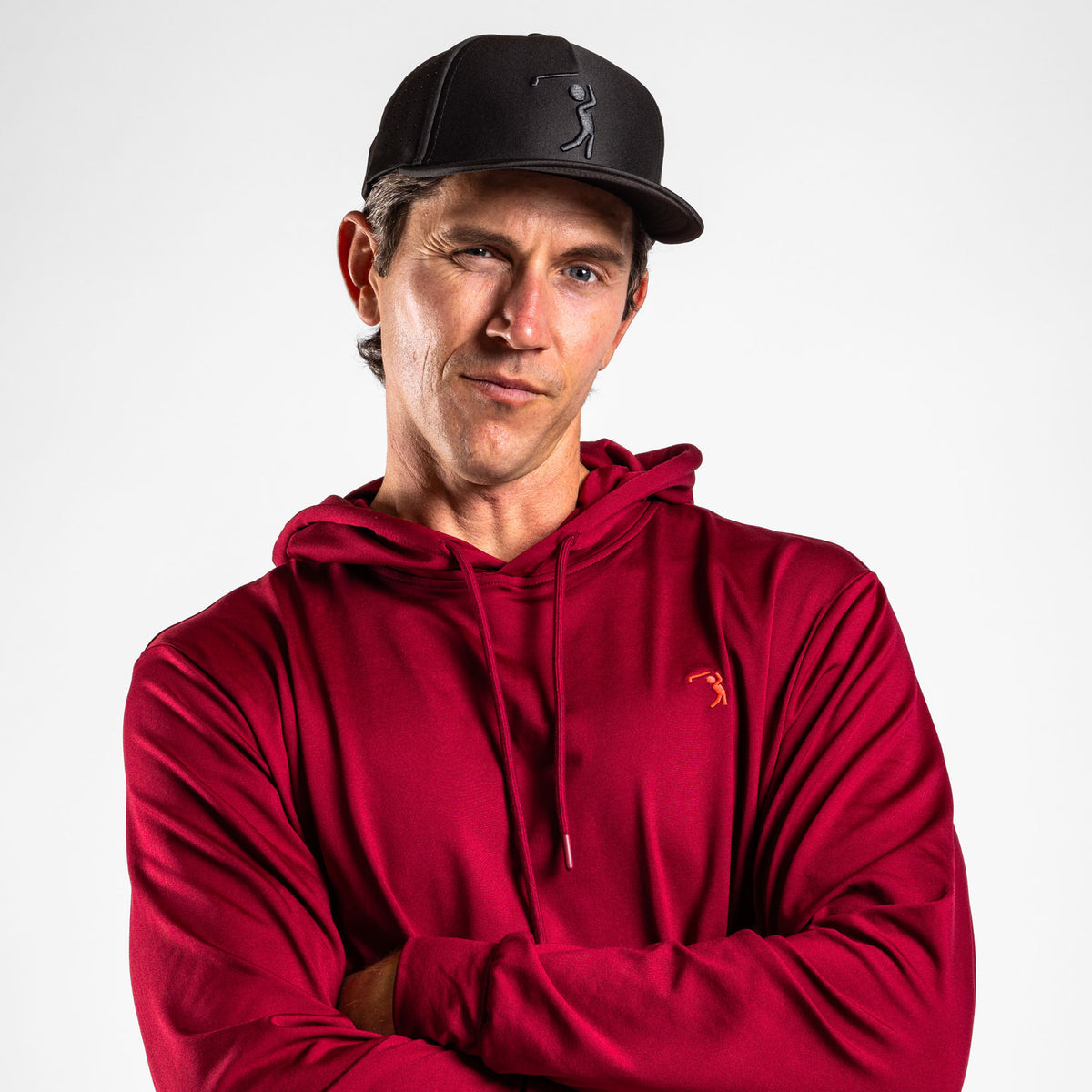 A man wearing the Burgundy - Hoodie and a black baseball cap with a golfer logo stands against a plain white background, looking at the camera and winking. With one arm crossed and the other hand touching his chin in a playful, confident pose, he perfectly pairs casual style with performance-ready athletic fit shorts featuring moisture-wicking comfort.