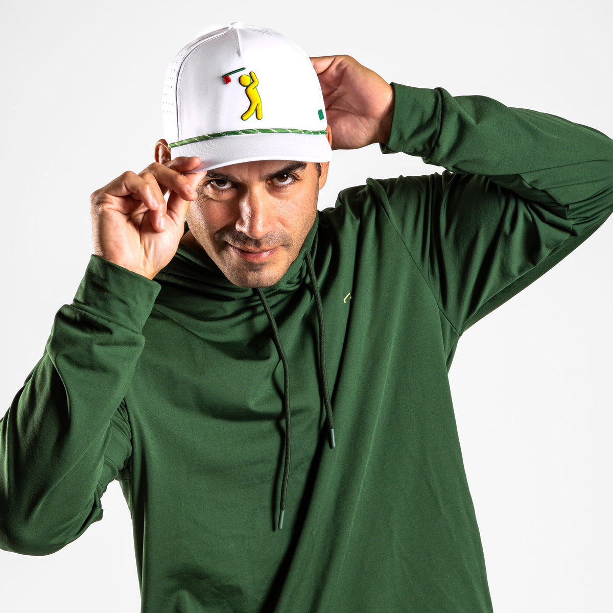 A man wearing the Forest Green - Hoodie tilts a white cap with a yellow golfer graphic, looking at the camera with a slight smile. His right hand adjusts the brim of the cap, while his moisture-wicking athletic shorts complete his sporty look against a plain white background.