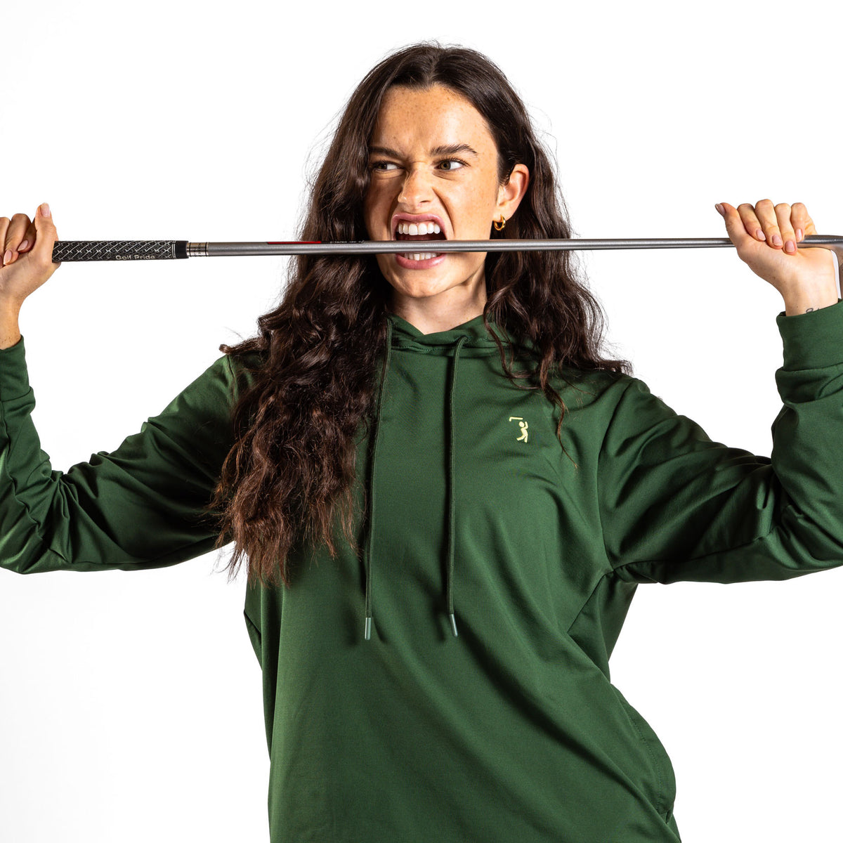 A woman wearing the Forest Green - Hoodie and athletic shorts playfully bites a golf club held horizontally in front of her mouth. She flashes a playful, intense smile, her long hair cascading over her shoulders against a plain white background. Her stretchy, moisture-wicking shorts enhance the sporty vibe of the scene.
