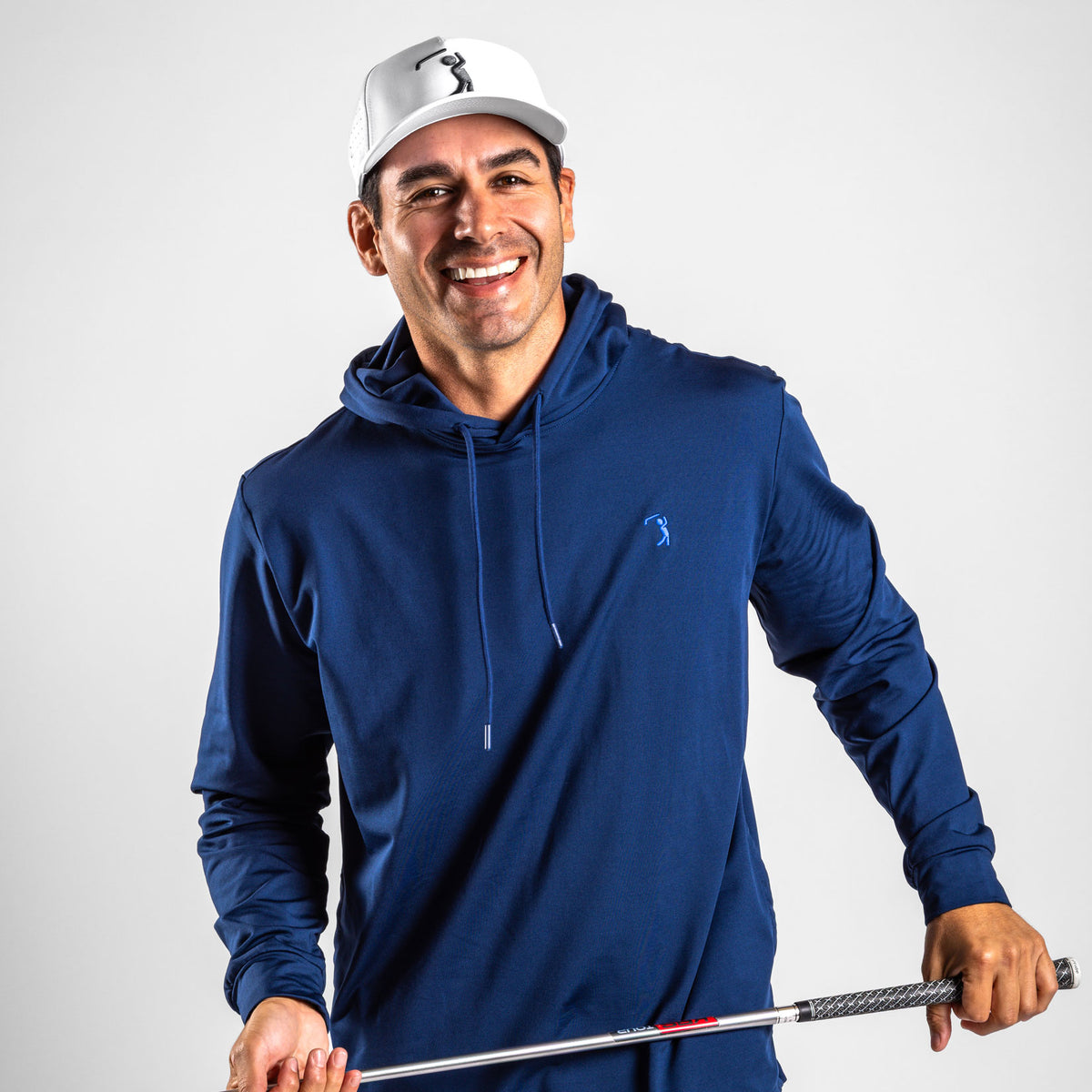 A smiling, dark-haired man wears the Navy - Hoodie and a white golf cap with a logo as he stands confidently against a plain, light grey background. The Navy - Hoodie features a small, light blue golfer silhouette logo on the chest and is crafted from moisture-wicking fabric to keep him comfortable on the course. Holding a golf club horizontally in front of him, he’s ready for his next swing.