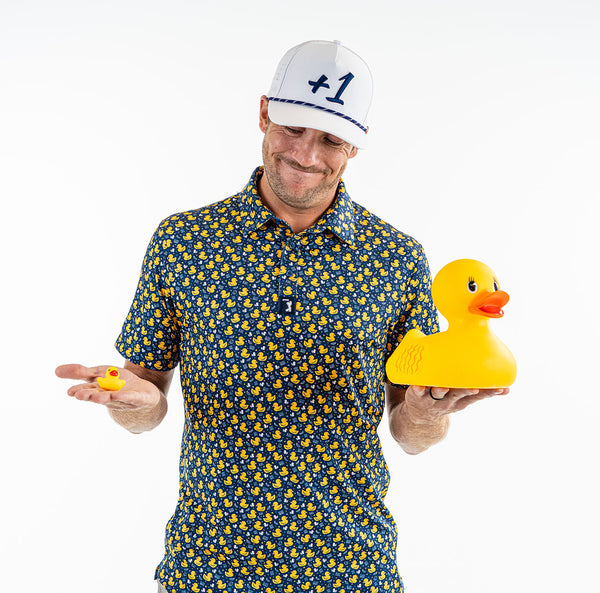 A smiling man wears the Big Duck Energy - Polo, a blue moisture-wicking shirt with yellow rubber duck print, and a white +1 cap. He holds small and large rubber ducks while standing against a plain white background.