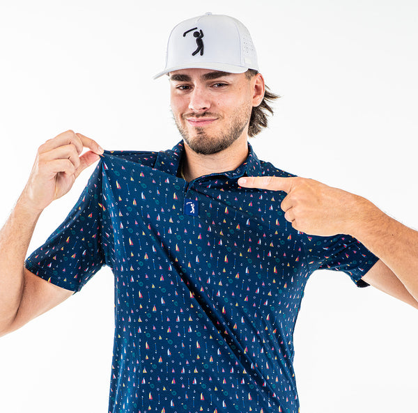 A man wearing the Boats and Hose - Polo—a navy athletic fit shirt with a small colorful sailboat pattern—stretches its collar and points at it, smiling slightly. The polo features moisture-wicking fabric. The background is plain white.