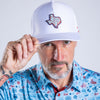 A man wearing a white trucker hat with a 'Bust the Texas' patch and a light blue patterned button-down shirt, posing against a light gray background.