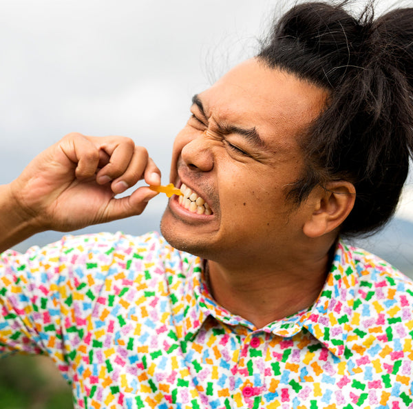 A man with dark hair in a topknot, wearing a colorful Gummy Bears - Polo, bites a gummy candy and squints as if it's tough to chew, set against a blurred outdoor background.